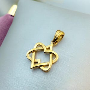 Op de afbeelding: Een goudkleurige hanger met een verweven hart- en sterontwerp. De hanger heeft een gepolijste afwerking en een kleine lus voor een ketting. De sieraden worden gepresenteerd op een lichtgekleurd oppervlak.