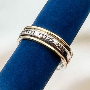 May include: A silver and gold spinning ring with Hebrew text engraved on the band. The ring is on a blue surface.