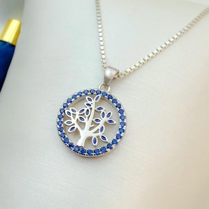 May include: Silver-toned necklace featuring a circular pendant with a tree design. The tree is silver, surrounded by a ring of blue gemstones. The necklace has a delicate chain and is set against a white background.