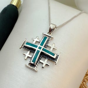 May include: A silver necklace with a turquoise Jerusalem cross pendant. The cross is detailed with a rope-like design and has a turquoise inlay.