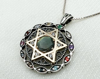 Op de afbeelding: Een zilveren en gouden ketting met een hanger in de vorm van een Davidster met een groene edelsteen in het midden, omringd door kleinere witte edelstenen. De hanger is omlijst door een cirkel van kleinere edelstenen in verschillende kleuren.