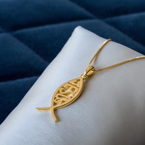 Könnte beinhalten: Eine goldfarbene Anhängerkette mit dem Ichthys-Symbol, wobei das Wort "JESUS" in der Fischform ausgeschnitten ist. Die Kette wird auf einer weißen Oberfläche vor einem dunkelblauen Hintergrund präsentiert.