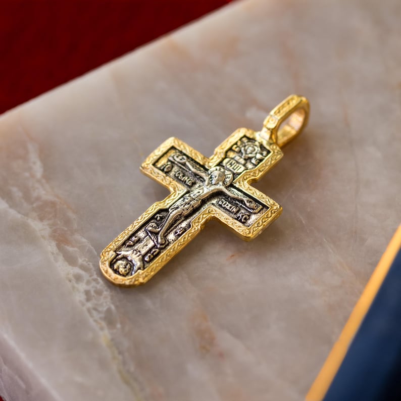 Puede incluir: Un colgante de cruz de metal en tono dorado con un relieve central en tono plateado de una figura crucificada. La cruz tiene un borde texturizado y ornamentado e incluye peque&ntilde;os detalles de texto cir&iacute;lico grabados en la secci&oacute;n central.