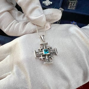 Pu&ograve; includere: Una collana con un ciondolo d'argento a forma di croce di Gerusalemme con una pietra preziosa blu al centro.