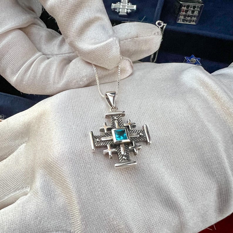 Pu&ograve; includere: Una collana con un ciondolo d'argento a forma di croce di Gerusalemme con una pietra preziosa blu al centro.