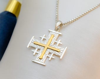 May include: Silver-tone necklace with a Jerusalem cross pendant. The pendant features a gold-colored cross overlaid on a silver cross design. The necklace is displayed on a white surface, with a blurred blue and gold object in the background.