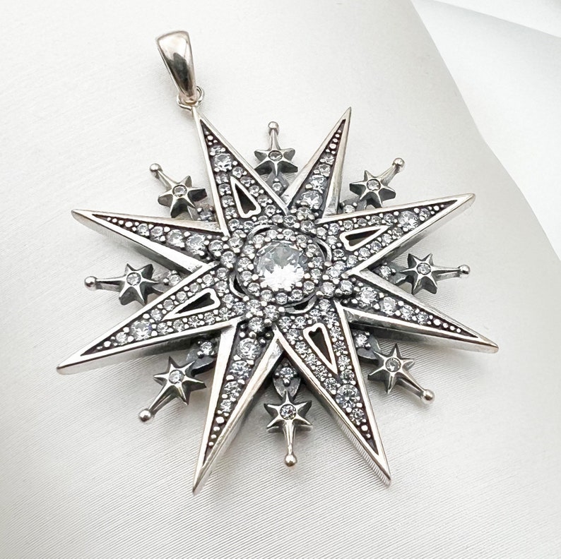 May include: A silver pendant in the shape of a star with a large clear crystal in the center. The star is surrounded by smaller crystals.