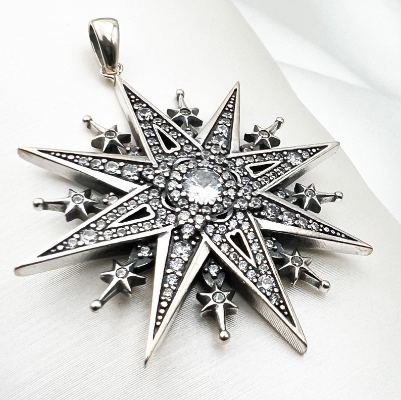 May include: A silver pendant in the shape of a large, eight-pointed star with smaller stars around the perimeter. The star is covered in clear, sparkling rhinestones.