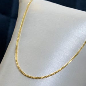 May include: A thin, gold-colored wheat chain necklace featuring a tightly woven, braided link pattern. The chain has a polished, reflective metallic finish and is draped in a smooth, curved U-shape, showcasing the intricate texture of the links.