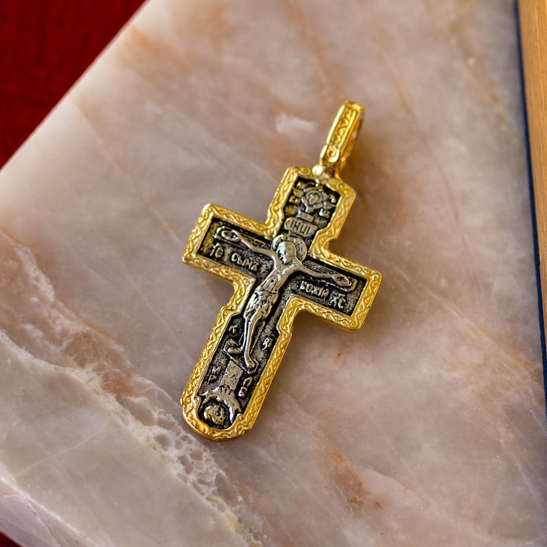 Puede incluir: Un colgante de cruz de metal dorado con un relieve central plateado de una figura crucificada. La cruz tiene un borde intrincado e incluye grabados en cir&iacute;lico. Una peque&ntilde;a anilla est&aacute; unida en la parte superior para colgar.