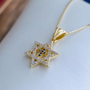 Könnte beinhalten: Eine goldfarbene Halskette mit einem Davidstern-Anhänger und einem Kreuz in der Mitte. Der Stern ist mit kleinen, klaren Kristallen besetzt, und das Kreuz besteht aus schwarzen Steinen. Die Halskette liegt auf einer weißen Oberfläche.