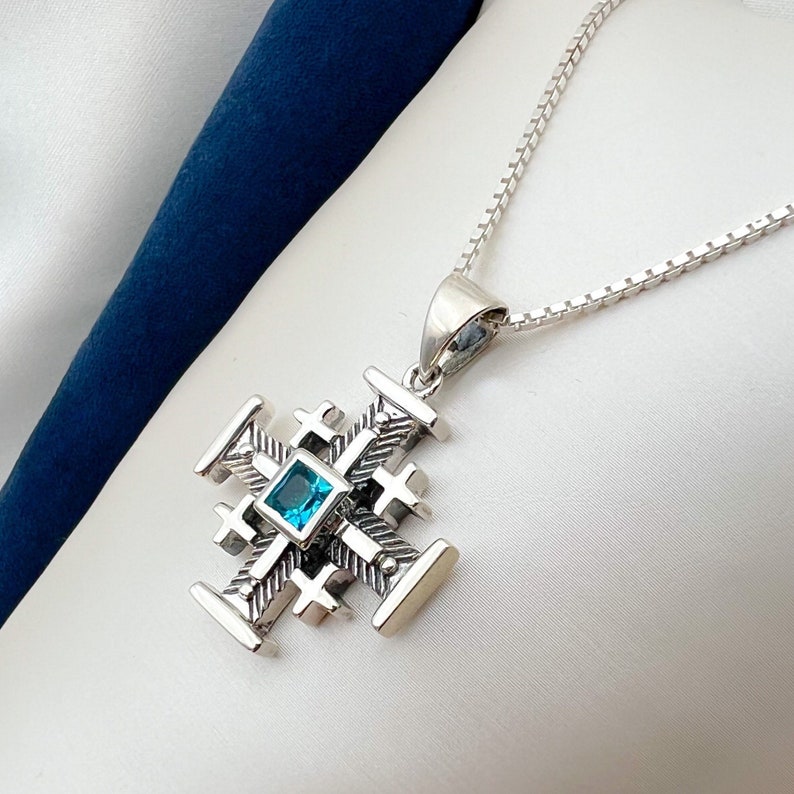 Pu&ograve; includere: Una collana con catena d'argento con un ciondolo d'argento a forma di croce. La croce ha una pietra preziosa blu al centro.
