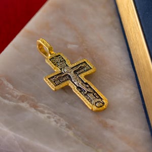 Puede incluir: Un colgante de cruz de metal dorado con un relieve central plateado de un crucifijo, detalles grabados intrincados y letras cir&iacute;licas. La cruz presenta un borde texturizado y una peque&ntilde;a anilla integrada en la parte superior.