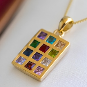 Op de afbeelding: Een goudkleurige ketting met een rechthoekige hanger met een raster van kleurrijke vierkante edelstenen. De edelstenen omvatten tinten rood, blauw, groen, paars en geel. De ketting is ook goudkleurig.