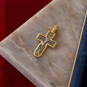 Puede incluir: Un colgante bicolor con forma de cruz, que presenta un borde dorado texturizado y una incrustaci&oacute;n central plateada que representa una figura en una cruz con letras cir&iacute;licas grabadas y detalles de iconograf&iacute;a religiosa.
