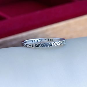 May include: A silver ring with an engraved pattern and Hebrew text. The ring has a thin band with a repeating swirl design. The ring is displayed on a white surface with a blurred red background.