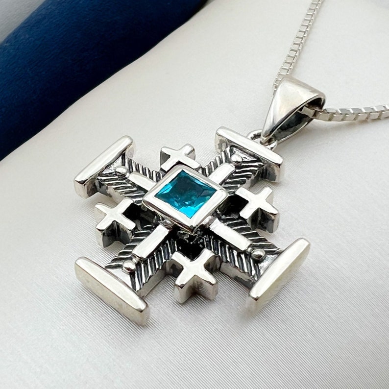 Pu&ograve; includere: Una collana con un ciondolo in argento a forma di croce con una pietra preziosa blu al centro.