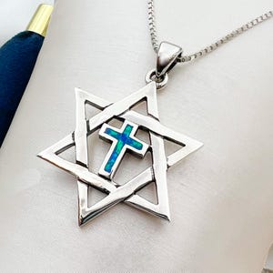 Puede incluir: Un collar con colgante plateado con un diseño de Estrella de David con una cruz en el centro. La cruz tiene una incrustación de ópalo azul y verde. El collar está sobre un fondo blanco.