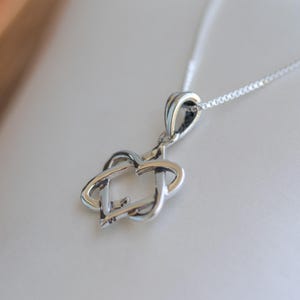 May include: A silver pendant necklace featuring an intertwined heart and key design. The pendant hangs from a delicate chain, resting on a white surface. The necklace is a piece of jewelry.
