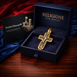 Puede incluir: Un colgante de crucifijo en tonos oro y plata oscura con una figura detallada de Jes&uacute;s, dentro de una caja azul marino con la etiqueta RELIGIONE FINE JEWELRY. Una tarjeta de certificado de garant&iacute;a negra de Jerusalem Diamonds est&aacute; al lado.