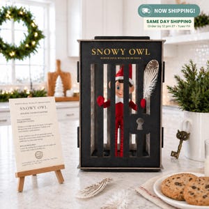 May include: A black wooden cage with the text SNOWY OWL NORTH POLE WILDLIFE RESERVE containing a small elf doll in a red outfit. Beside it stands a sign titled SNOWY OWL HOLIDAY INCIDENT REPORT on a wooden easel, with a key nearby.