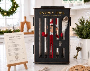 May include: A black wooden cage with the text SNOWY OWL NORTH POLE WILDLIFE RESERVE containing a small elf doll in a red outfit. Beside it stands a sign titled SNOWY OWL HOLIDAY INCIDENT REPORT on a wooden easel, with a key nearby.