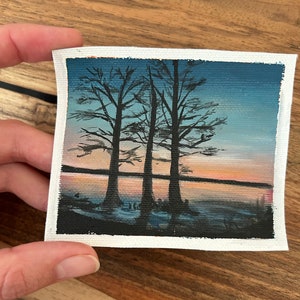 Puede incluir: Pequeña pintura de paisaje que representa tres árboles silueteados contra un atardecer. La obra de arte utiliza colores azul, naranja y negro, montada sobre un lienzo con un borde blanco, creando una escena tranquila.