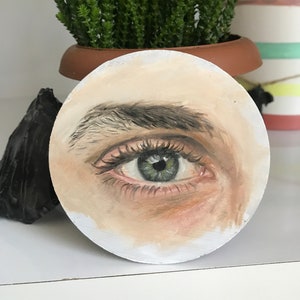 Puede incluir: Obra de arte circular pintada que representa un ojo humano detallado. El iris es verde, con pesta&ntilde;as y cejas visibles, sobre un fondo de piel clara y un fondo blanco. Una maceta con una planta es visible en el fondo.
