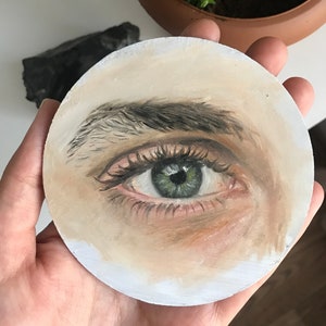 Puede incluir: Una obra de arte redonda, pintada a mano, que presenta un ojo detallado con iris verde, pesta&ntilde;as oscuras y un tono de piel claro. El ojo es el punto focal, con sombreado y reflejos sutiles. La obra de arte se sostiene en una mano, con una planta y una roca oscura en el fondo.