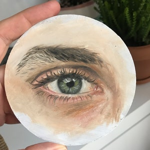 Puede incluir: Una obra de arte redonda pintada que presenta una representaci&oacute;n detallada de un ojo humano. El ojo es verde con largas pesta&ntilde;as y una ceja definida. La obra de arte est&aacute; pintada sobre una superficie circular blanquecina.