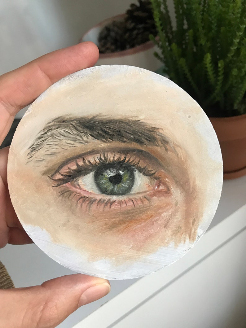 Puede incluir: Una obra de arte redonda pintada que presenta una representaci&oacute;n detallada de un ojo humano. El ojo es verde con largas pesta&ntilde;as y una ceja definida. La obra de arte est&aacute; pintada sobre una superficie circular blanquecina.