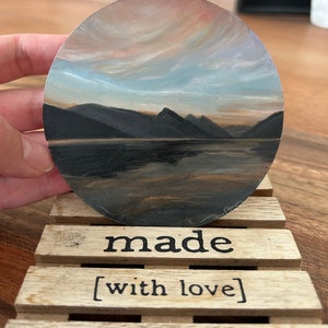 Puede incluir: Pintura redonda de un paisaje montañoso con un lago, con un cielo de atardecer con tonos rosas y azules. La pintura está sobre un posavasos de madera que dice "made [with love]" en texto negro.