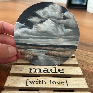 Puede incluir: Pintura redonda de un paisaje con un cielo nublado y una masa de agua. La pintura se sostiene sobre una paleta de madera que dice "made [with love]" en texto negro.