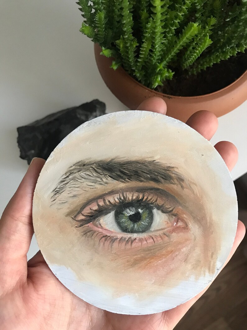 Puede incluir: Una obra de arte redonda, pintada a mano, que presenta un ojo detallado con iris verde, pesta&ntilde;as oscuras y un tono de piel claro. El ojo es el punto focal, con sombreado y reflejos sutiles. La obra de arte se sostiene en una mano, con una planta y una roca oscura en el fondo.