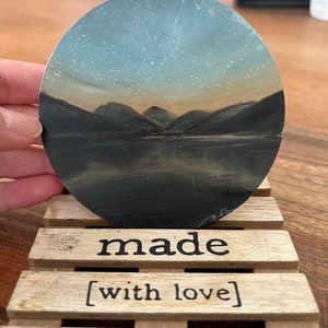 Puede incluir: Una pintura redonda representa un paisaje sereno con montañas, agua y un cielo estrellado. La pintura descansa sobre una pequeña paleta de madera que dice "made [with love]" en letras negras. La obra de arte es sostenida por una mano.