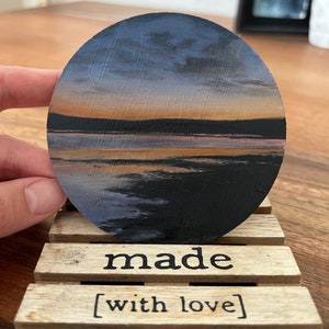 Puede incluir: Una pequeña pintura redonda de una puesta de sol sobre el agua, con tonos naranja, negro y azul. La pintura está sobre una paleta de madera que dice "made [with love]" en texto negro.