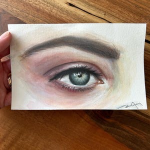 Puede incluir: Pintura a la acuarela de un ojo humano con una representación detallada del iris, las pestañas y la ceja. El ojo es de color azul verdoso claro, rodeado de tonos rosas y morados. La obra de arte está sobre un trozo de papel rectangular.