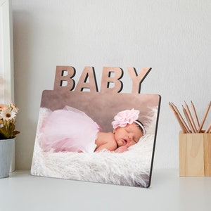 May include: A wooden photo frame with the word "BABY" cut out at the top. The frame holds a photo of a sleeping infant wearing a pink tutu and a flower headband. The baby is lying on a fluffy white surface.