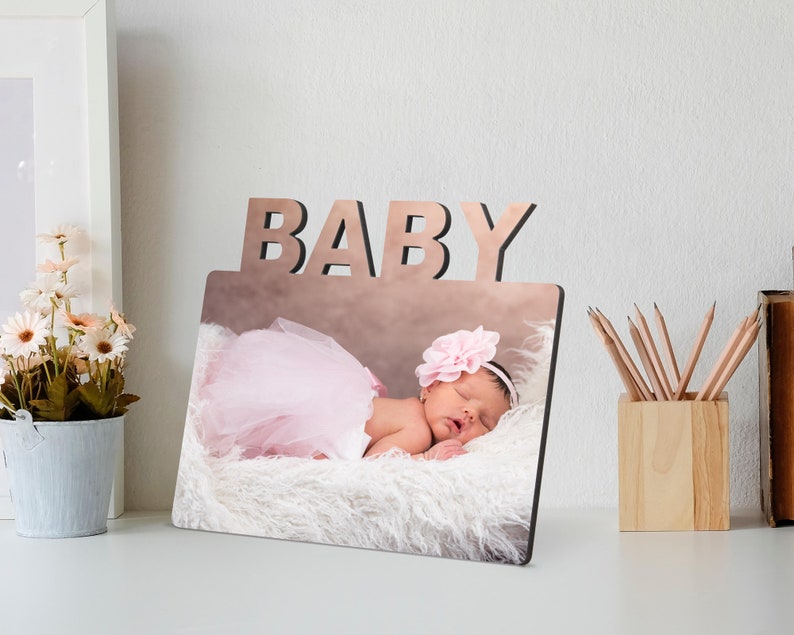 May include: A wooden photo frame with the word "BABY" cut out at the top. The frame holds a photo of a sleeping infant wearing a pink tutu and a flower headband. The baby is lying on a fluffy white surface.