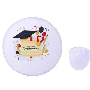 May include: A white graduation button with a black mortarboard, diploma, and the words "Happy Graduation." The design includes red, gold, and black accents. A separate, white, shield-shaped object is next to the button.