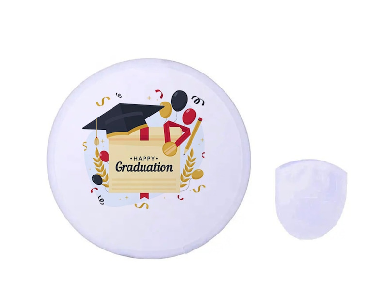 May include: A white graduation button with a black mortarboard, diploma, and the words "Happy Graduation." The design includes red, gold, and black accents. A separate, white, shield-shaped object is next to the button.