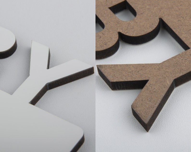 May include: Close-up of wooden letters, one white and one brown, with a dark edge. The letters are cut from a thick material, creating a 3D effect. The background is a plain white surface.