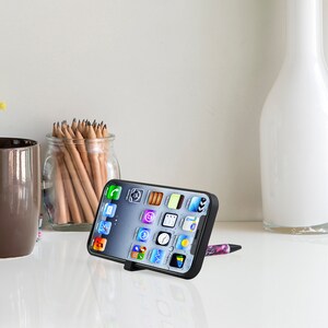 May include: A black phone stand with a phone propped up on it. The phone is displaying a variety of apps on the screen. The stand is on a white surface.
