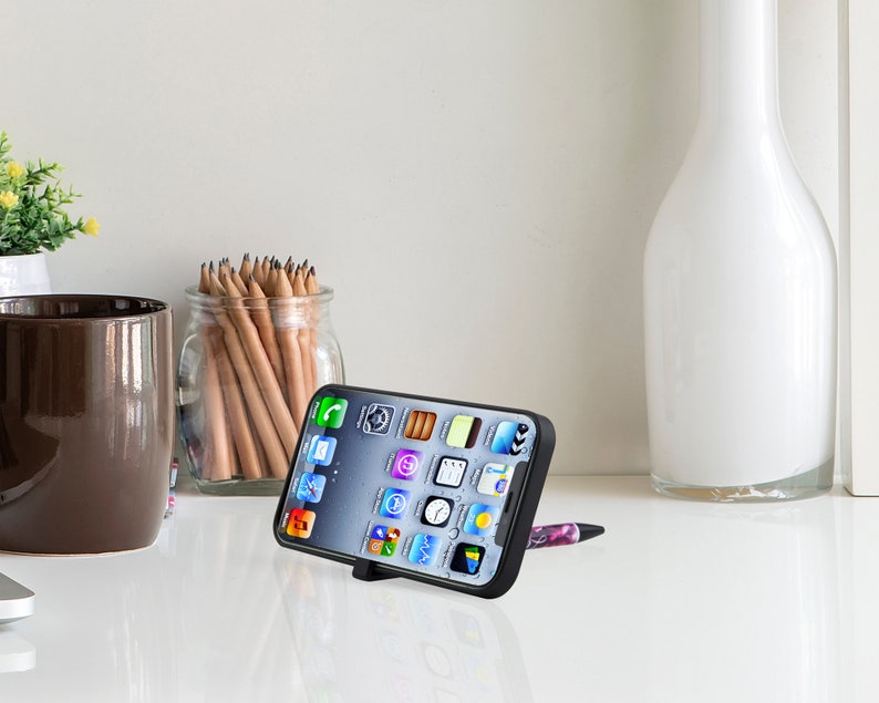 May include: A black phone stand with a phone propped up on it. The phone is displaying a variety of apps on the screen. The stand is on a white surface.
