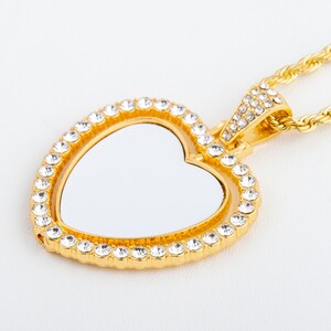 May include: A gold heart-shaped pendant necklace with a clear crystal encrusted border. The pendant has a blank space in the centre for a photo or inscription. The necklace is attached to a gold rope chain.