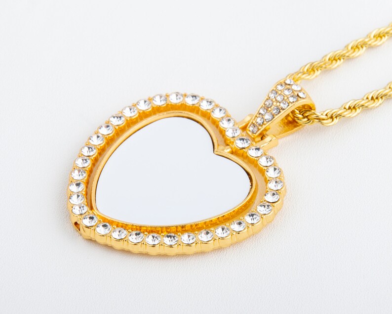 May include: A gold heart-shaped pendant necklace with a clear crystal encrusted border. The pendant has a blank space in the centre for a photo or inscription. The necklace is attached to a gold rope chain.