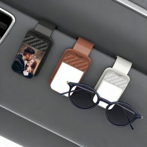 May include: Three car visor clips in black, brown, and white. The black clip displays a photo of a couple. A pair of dark blue sunglasses rests below the clips. The clips have a textured design.
