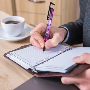 May include: A person writing in a brown leather bound planner with a purple and black pen that has a phone stand on the top. The pen has the text "Love you always" written on it.