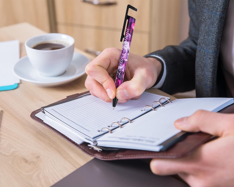 May include: A person writing in a brown leather bound planner with a purple and black pen that has a phone stand on the top. The pen has the text "Love you always" written on it.