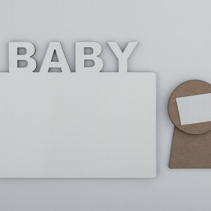 May include: A white wooden photo frame with the word "BABY" in capital letters above the frame. A separate, round, brown wooden piece with a white square is next to the frame.